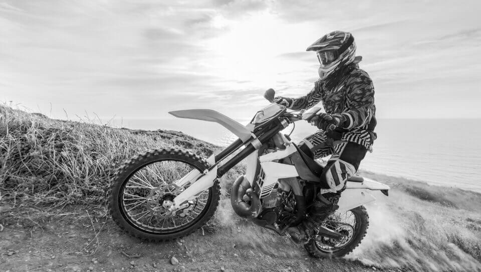 A motocross rider speeds on a dirt trail near a coastal cliff.